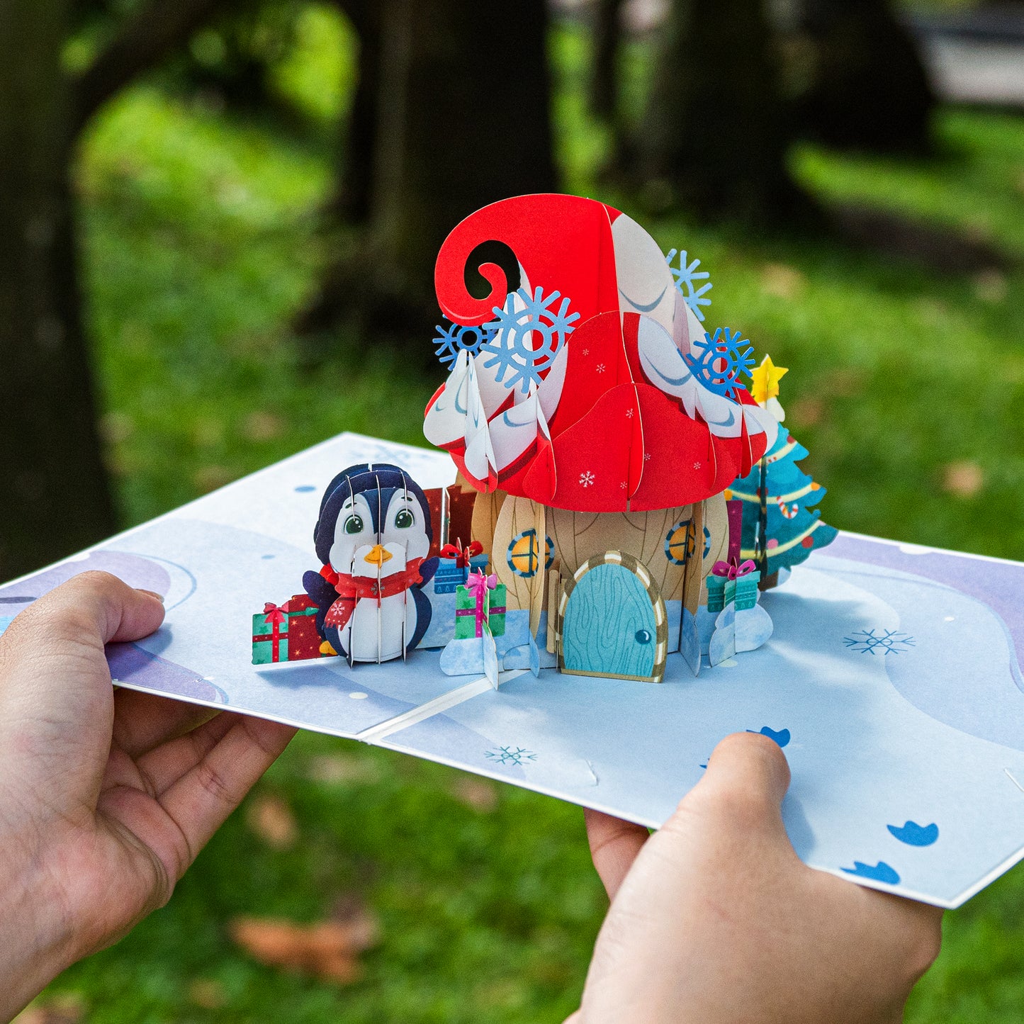 Hands holding a pop-up Christmas card with a mushroom house and penguin design.