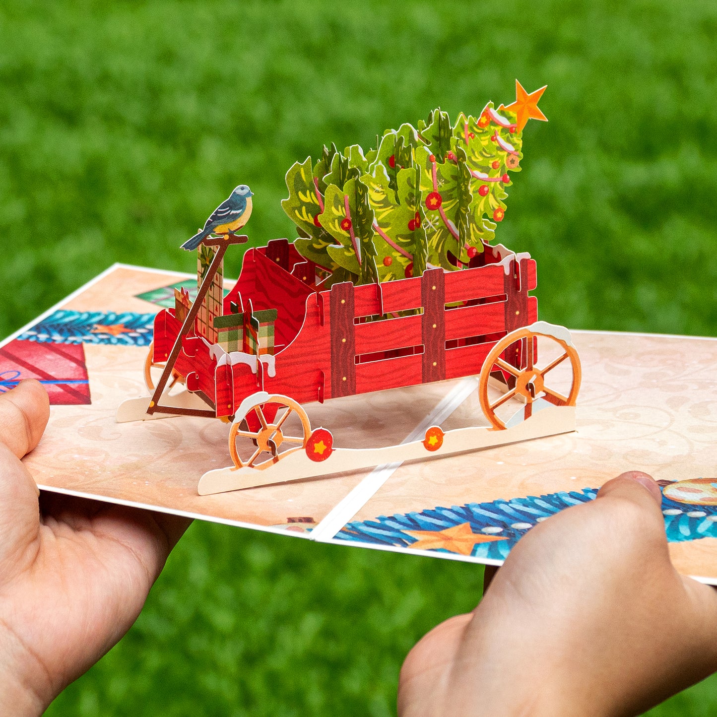 Hands holding a pop-up Christmas card with a red wagon and tree design.