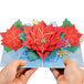 Hands holding a poinsettia pop-up Christmas card with intricate red flowers and gold accents.