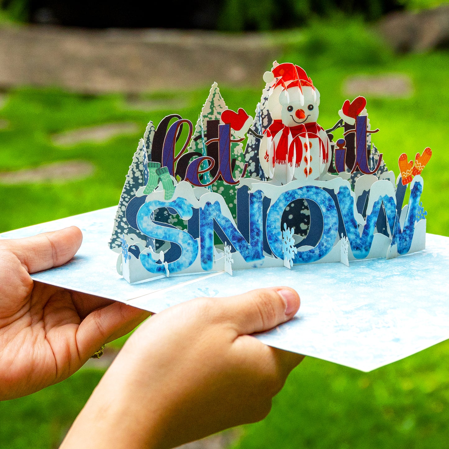 Hands holding Let It Snow pop-up card with vibrant 3D winter scene.
