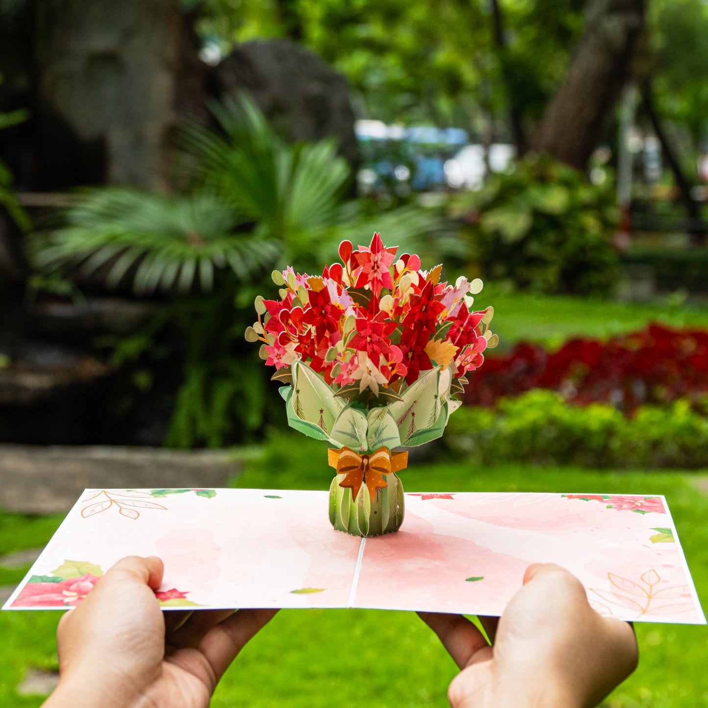 Hands holding poinsettia pop-up card in a lush outdoor setting.